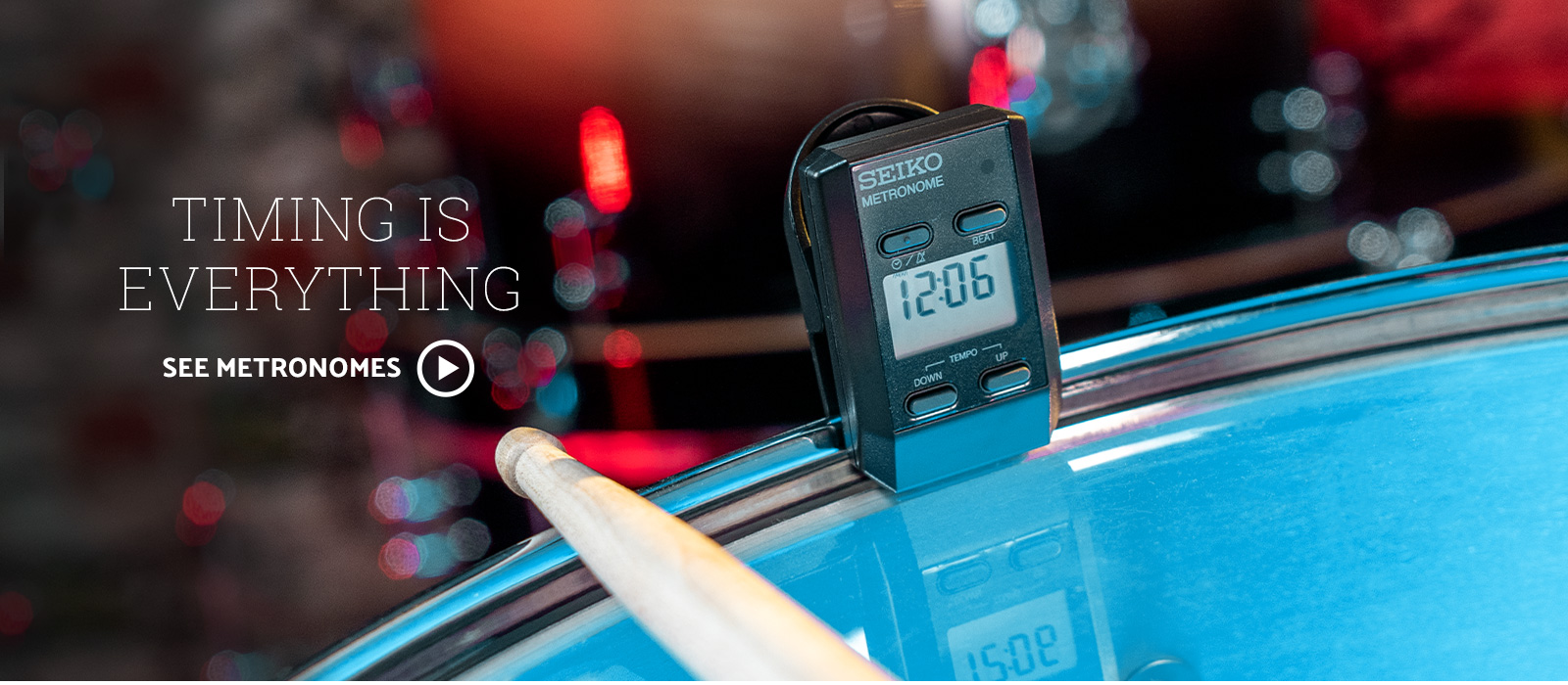 Seiko clip on metronome clipped to a drum kit. Image is lit with red and blue stage lighting. Text says Timing is Everything, click to see metronomes.