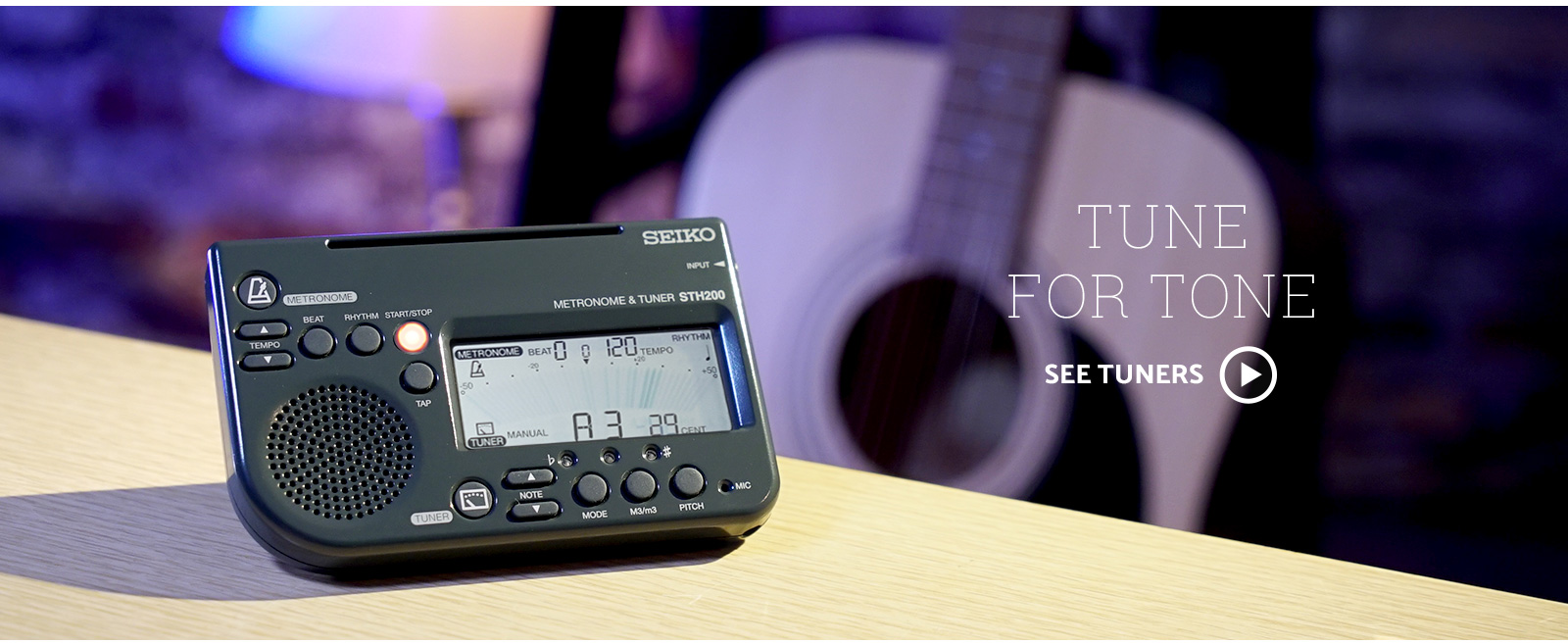 Seiko Tuner & Metronome combo sitting on the edge of a table. In the background is an acoustic guitar with purple stage lighting.